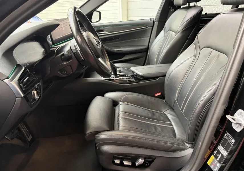 Vue intérieure côté conducteur de la BMW Série 5 Touring 530e noire avec sièges cuir et éclairage d'ambiance vert.