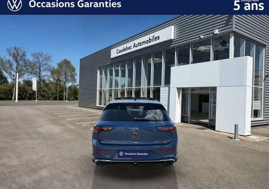 Vue arrière d'une Volkswagen Golf 1.5 eHybrid 272 DSG6 bleue métallisée, avec attelage rabattable, devant un bâtiment de concession.