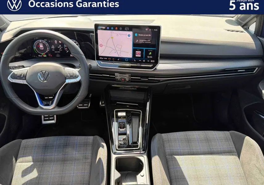 Vue intérieure avant de la Volkswagen Golf 1.5 eHybrid 2025, montrant le volant sport, l'écran tactile central et les sièges tissu à motif.