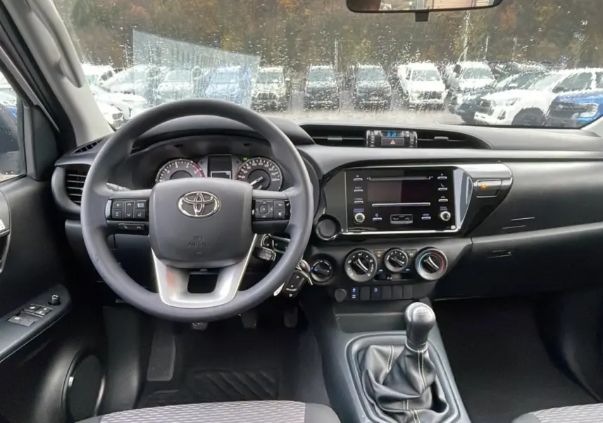 Vue intérieure avant du Toyota Hilux 2024 blanc, montrant le volant multifonction, tableau de bord et levier de vitesse manuel.