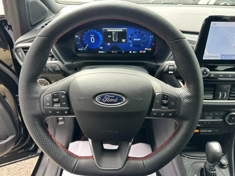 Vue intérieure centrée sur le volant perforé avec surpiqûres rouges du Ford Puma 2024, tableau de bord numérique en arrière-plan.