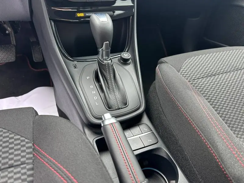 Gros plan sur la console centrale du Ford Puma 2024 noir, montrant le levier de vitesse automatique et le frein à main avec surpiqûres rouges.