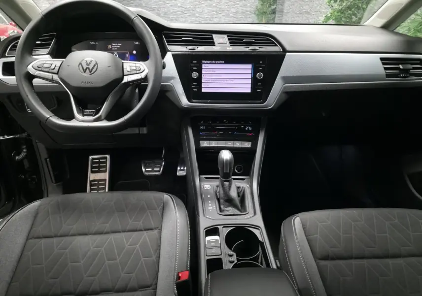 Vue intérieure avant du Volkswagen Touran 2025 noir, montrant le volant multifonction, l'écran tactile central et la console avec boîte auto.