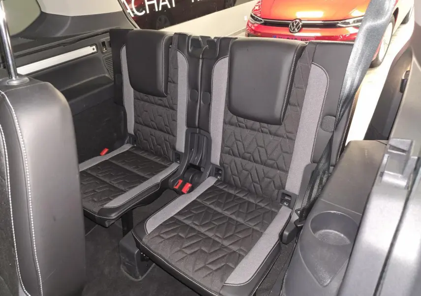 Vue intérieure sur la troisième rangée de sièges noirs et gris du Volkswagen Touran 2025, avec arrière d’un autre véhicule rouge visible.