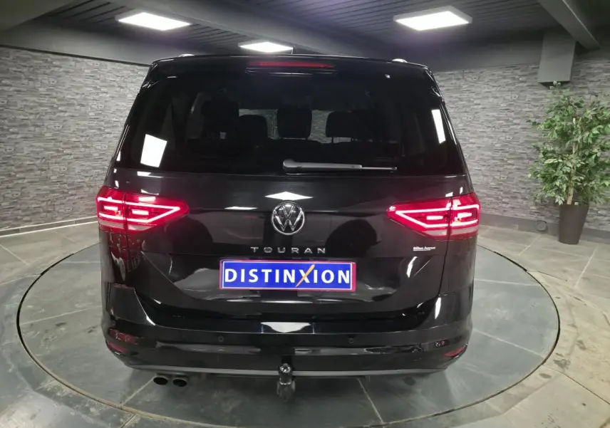 Vue arrière d'un Volkswagen Touran noir 2025 avec feux allumés et crochet d'attelage électrique visible.