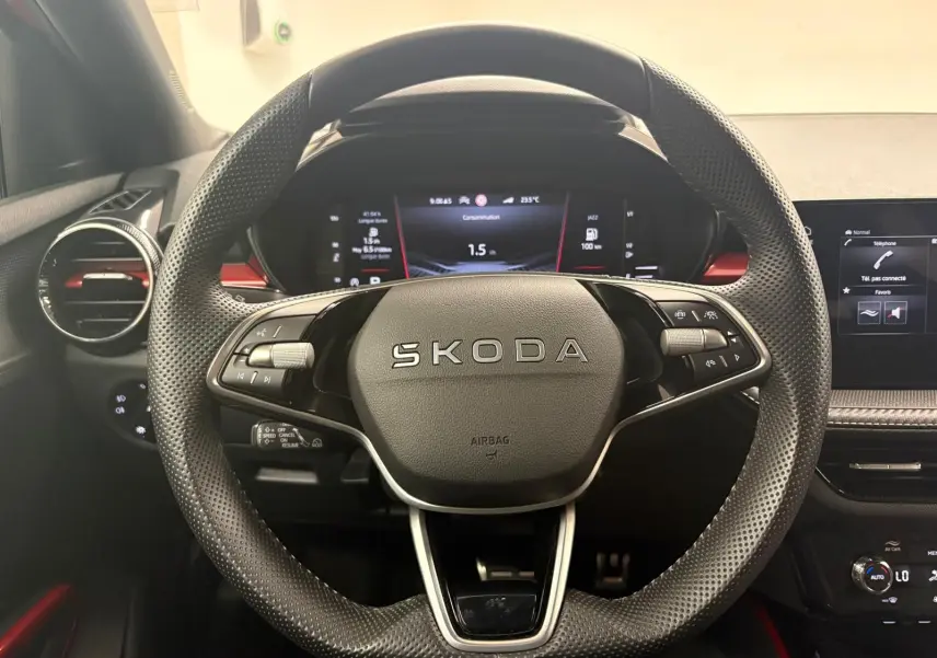 Vue rapprochée du volant perforé noir avec logo SKODA, tableau de bord numérique et détails rouges intérieurs visibles.