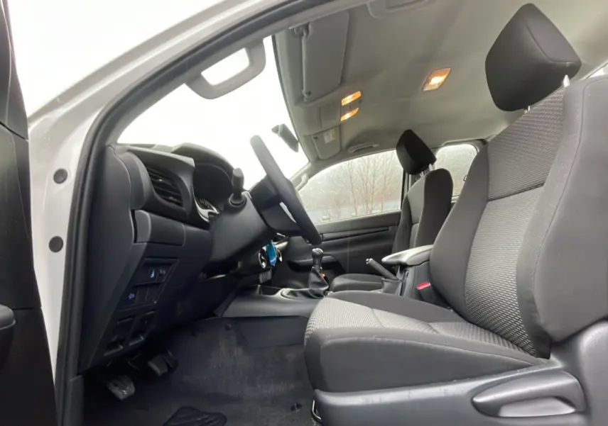 Intérieur du Toyota Hilux 4x4 2024 blanc, vue côté conducteur, sièges en tissu gris et tableau de bord noir.