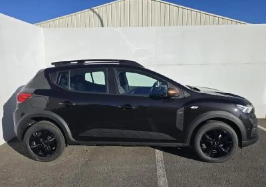 Profil côté gauche d'une Dacia Sandero III Stepway noir nacré avec barres de toit et jantes noires sur fond urbain.