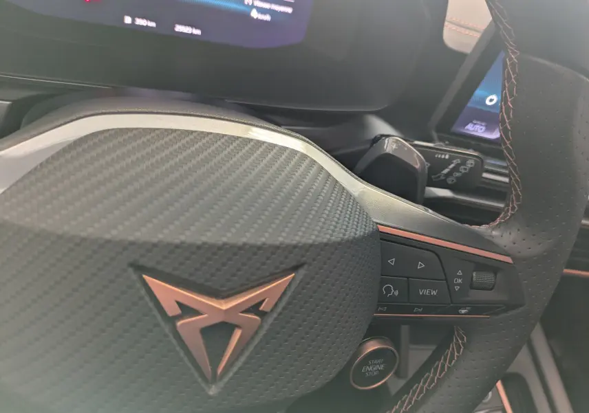 Gros plan sur le volant en cuir perforé avec logo CUPRA cuivré et commandes intégrées, tableau de bord numérique en arrière-plan.