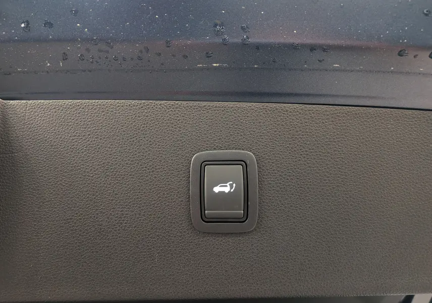 Gros plan sur le bouton d'ouverture électrique du hayon, sur fond intérieur noir du Dacia Bigster Bleu Indigo 2026.
