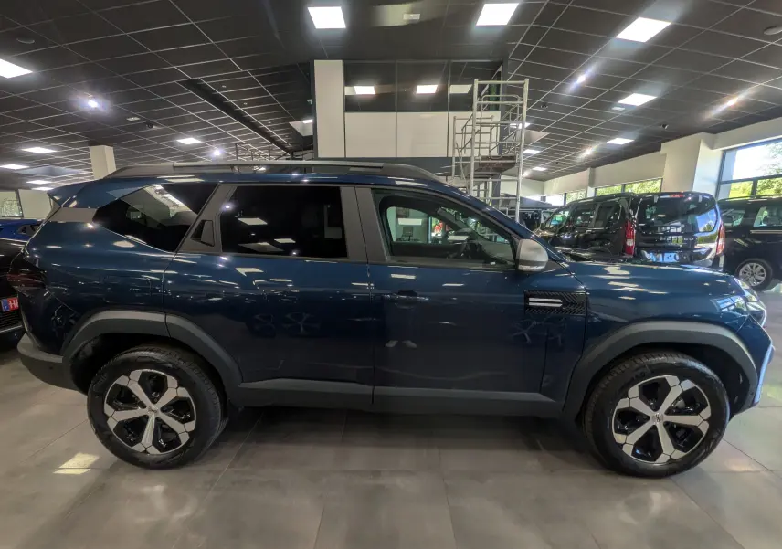 Vue de profil côté gauche du SUV Dacia Bigster bleu indigo avec jantes bicolores et toit noir en showroom.