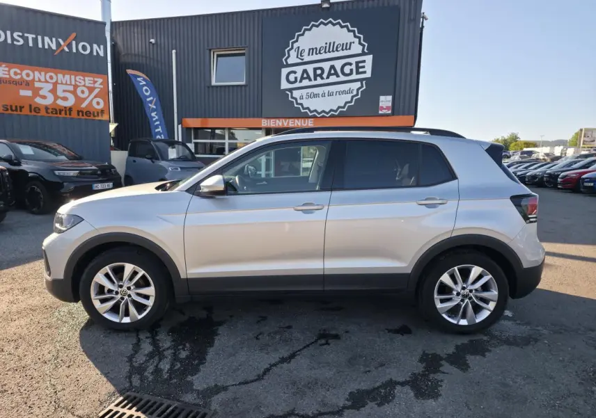 Profil côté gauche d'un Volkswagen T-Cross gris métallisé 2025 avec jantes alliage 17 pouces, stationné devant un garage.