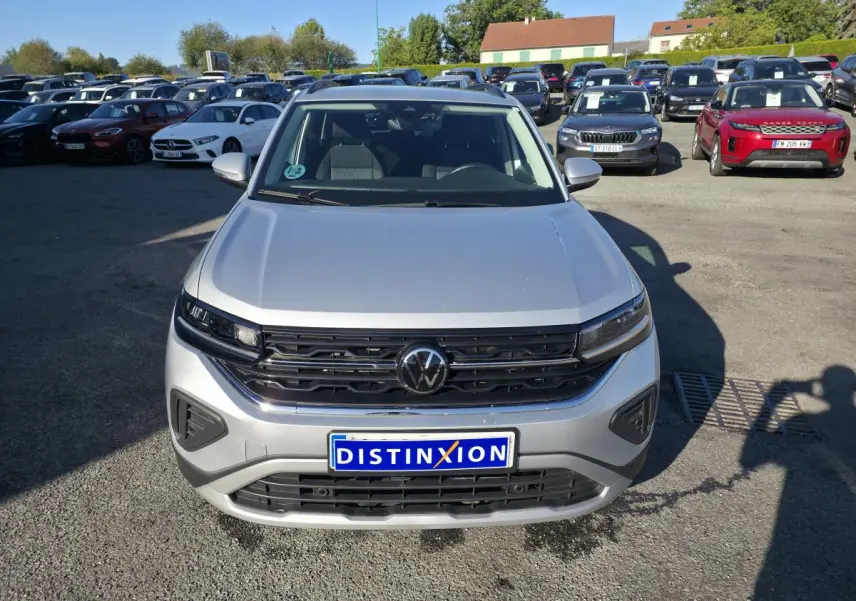 Vue avant d'un Volkswagen T-Cross gris métallisé 2025 avec calandre noire et logo VW central, sur parking extérieur.