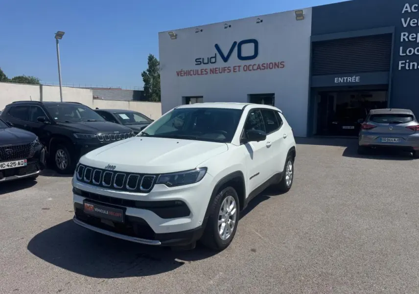 Jeep Compass blanc en 3/4 avant droit, stationné devant un bâtiment de concession sous un ciel bleu clair.