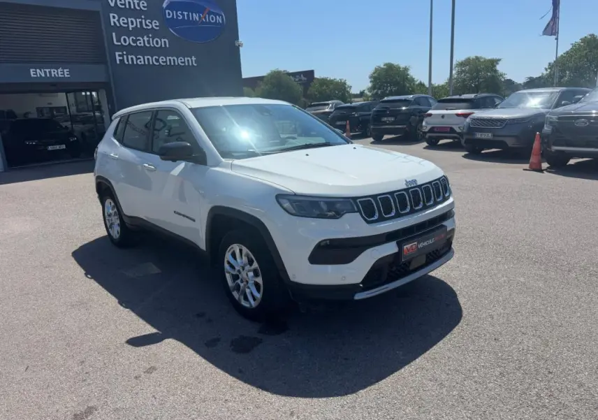 Jeep Compass blanc en vue 3/4 avant droit sur parking extérieur devant un bâtiment commercial.