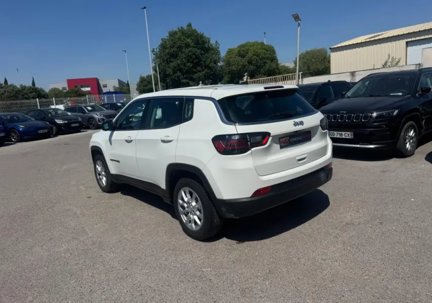 Jeep Compass blanc vu en 3/4 arrière droit sur parking, avec feux arrière sombres et jantes argentées.
