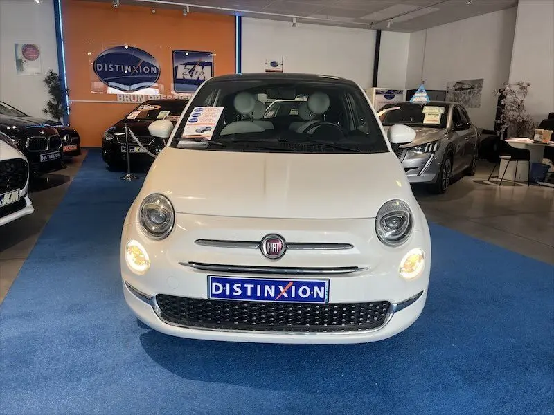 Vue frontale d'une Fiat 500 blanche avec phares allumés, exposée dans un showroom au sol bleu.