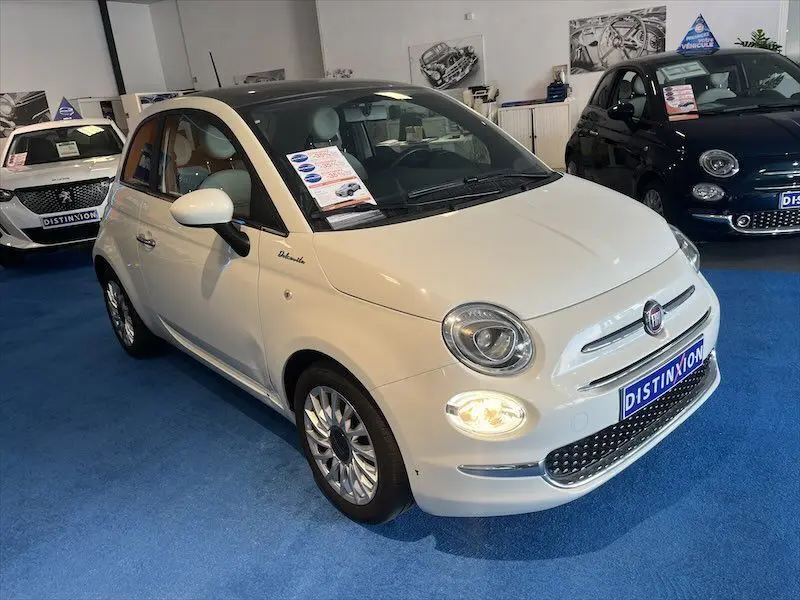 Fiat 500 blanche vue 3/4 avant droit en showroom avec phares allumés et toit noir contrasté.