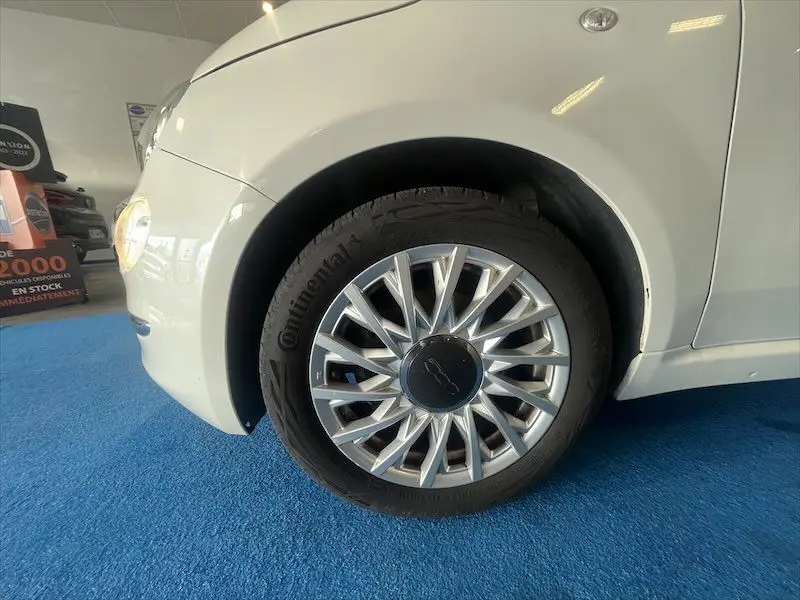 Gros plan sur la roue avant gauche d'une Fiat 500 blanche, mettant en valeur la jante argentée au design étoilé.