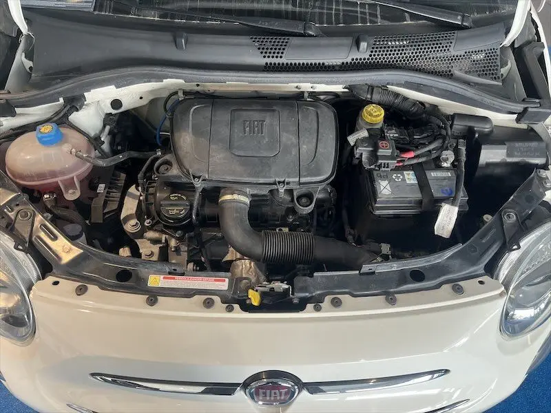 Vue plongeante sur le moteur d'une FIAT 500 blanche avec capot ouvert, montrant le compartiment moteur et la batterie.