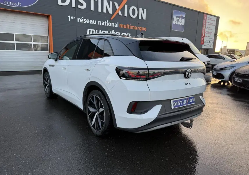 Vue 3/4 arrière droite d'un Volkswagen ID.4 GTX blanc glacier avec toit noir et crochet d'attelage sur parking humide.