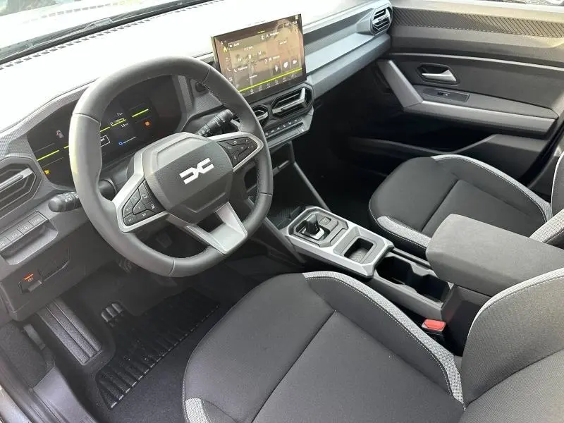 Vue intérieure côté conducteur du Dacia Duster 2025, montrant le volant, écran tactile et sièges noirs avec surpiqûres grises.