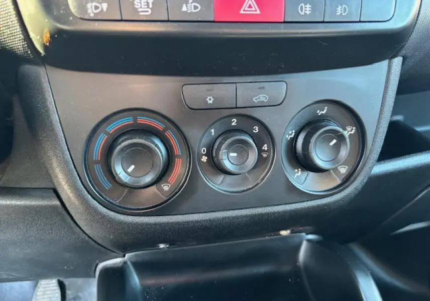Vue rapprochée des commandes de climatisation manuelle et des boutons de sécurité du tableau de bord d’un Opel Combo blanc de 2015.