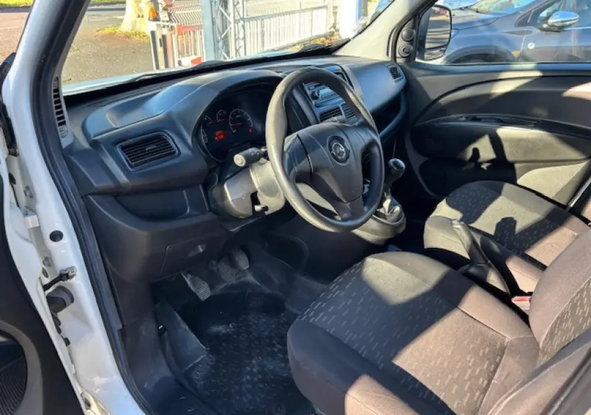 Vue intérieure côté conducteur de l'Opel Combo blanc, montrant le volant, tableau de bord et sièges avant en tissu noir.