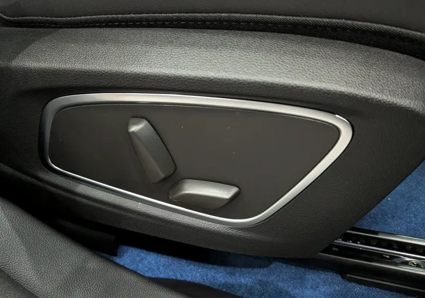 Gros plan sur les commandes électriques de réglage du siège côté gauche, intérieur noir avec moquette bleue visible.