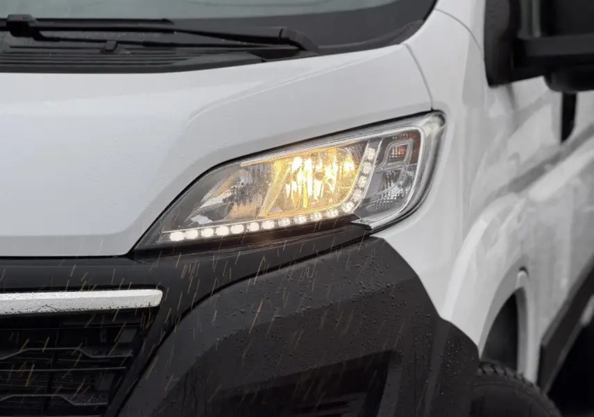 Gros plan sur le phare avant droit allumé d’un Opel Movano Fourgon blanc sous la pluie, avec pare-chocs noir.