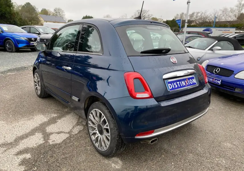 Vue 3/4 arrière droite d'une Fiat 500 bleu foncé avec toit panoramique, stationnée sur un parking extérieur.