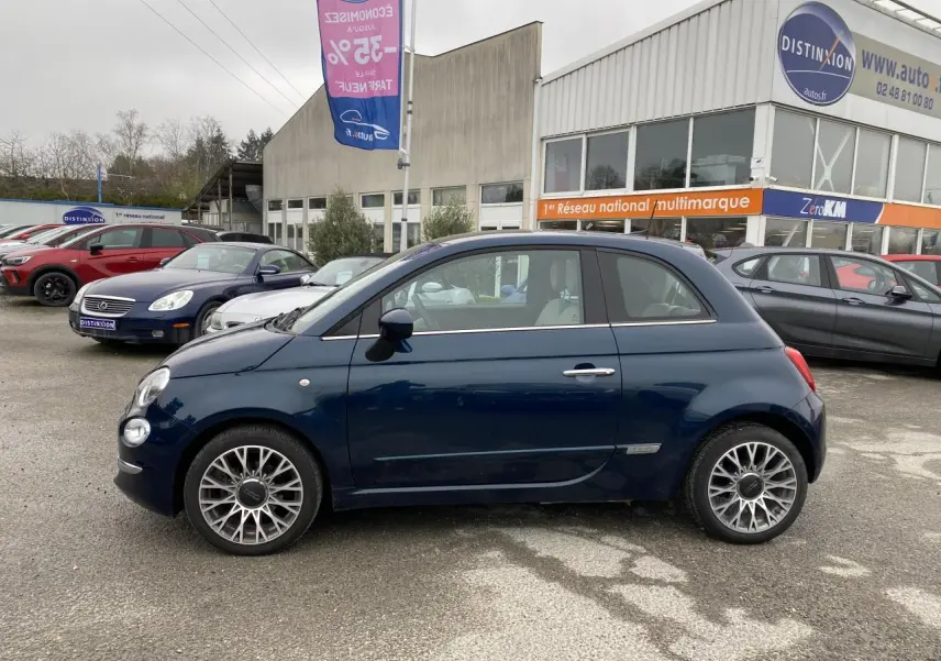 Vue de profil côté gauche d'une Fiat 500 bleu foncé 2019 avec toit panoramique, stationnée devant un concessionnaire.
