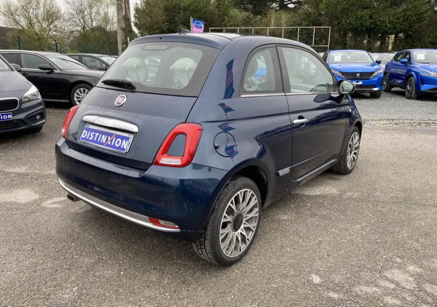 Vue 3/4 arrière droite d'une Fiat 500 bleu foncé avec toit panoramique, garée sur un parking extérieur.