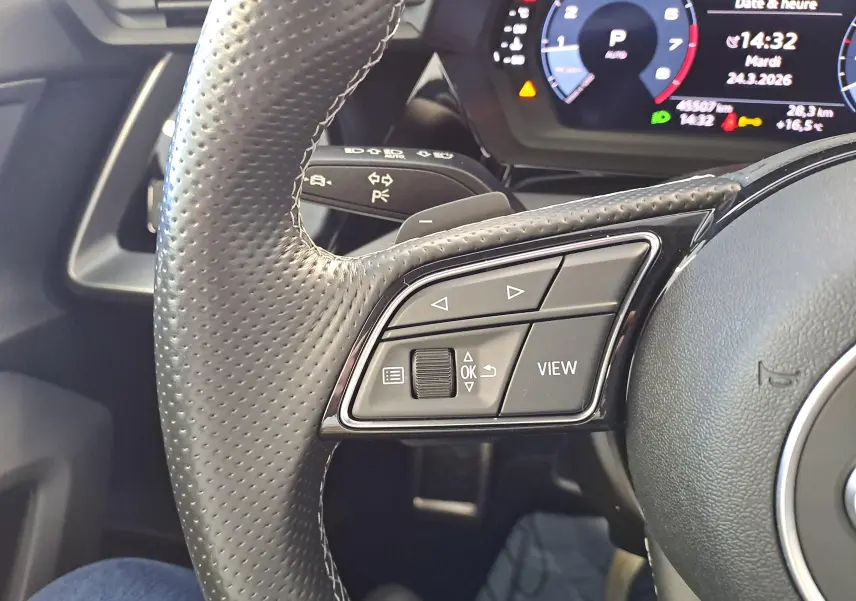 Gros plan sur le volant perforé côté gauche de l'Audi A3 Sportback noir mythic avec commandes multifonctions et tableau de bord digital.