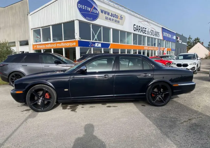 Vue latérale gauche d'une Jaguar XJ noire de 2006 avec jantes noires et étriers rouges, garée devant un garage multimarque.