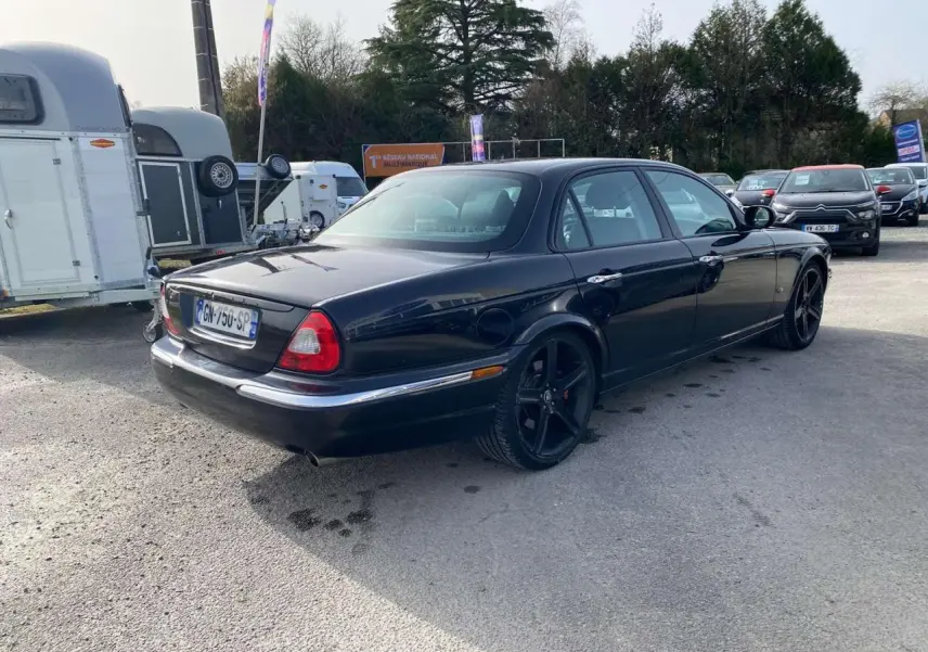 Jaguar XJ noire vue 3/4 arrière droit, avec jantes noires et feux arrière caractéristiques visibles.
