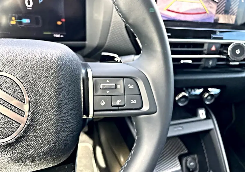 Gros plan sur le volant cuir noir et les commandes vocales du Citroën C4 X hybride 2025, avec écran tactile en arrière-plan.