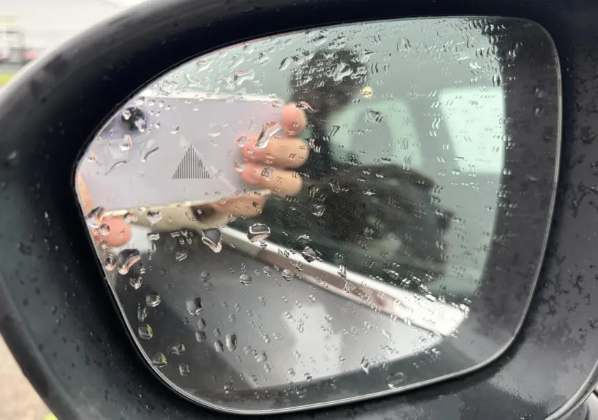 Rétroviseur extérieur droit du Nissan Qashqai 2025 gris foncé avec gouttes de pluie et témoin d'angle mort.