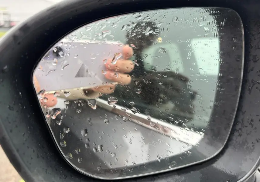 Rétroviseur extérieur droit noir avec gouttes d'eau sur un Nissan Qashqai gris foncé, reflet flou d'une main et d'un véhicule.