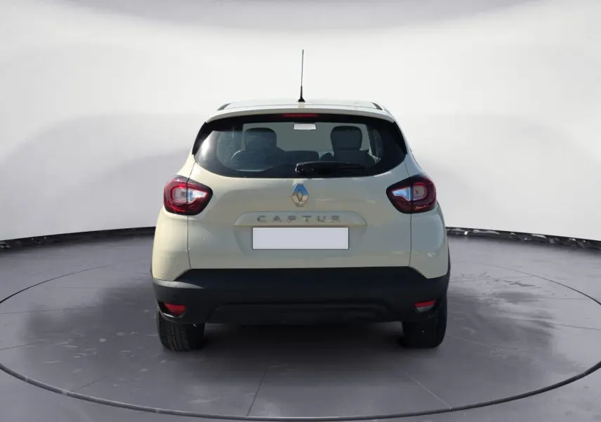 Vue 3/4 arrière droite d'un Renault Captur ivoire 2018 avec jantes alliage et protections noires sur un terrain extérieur.