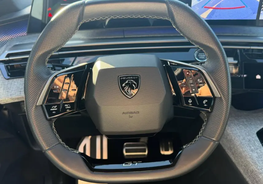 Vue rapprochée du volant cuir multifonction noir avec logo Peugeot et inscription GT, tableau de bord gris clair en arrière-plan.