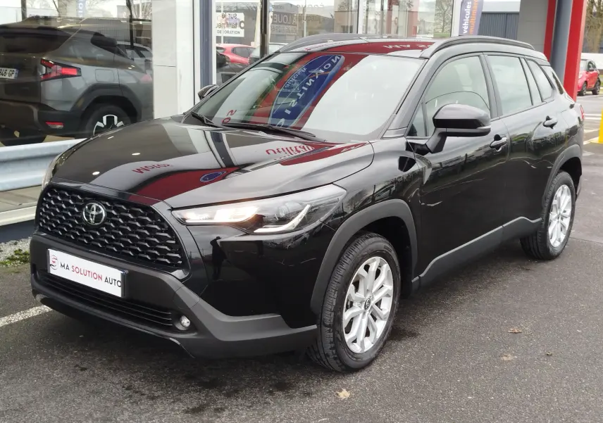 Toyota Corolla Cross noir intense vue 3/4 avant droit, avec calandre noire et jantes argentées, stationnée devant un showroom.