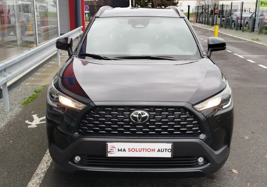Vue frontale d'un Toyota Corolla Cross noir intense avec calandre noire et phares allumés, stationné devant un bâtiment.