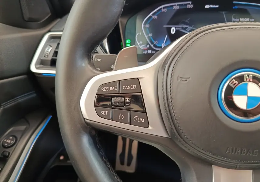 Gros plan sur le volant cuir noir de la BMW Série 3 320e hybride, avec commandes de régulateur et tableau de bord numérique.