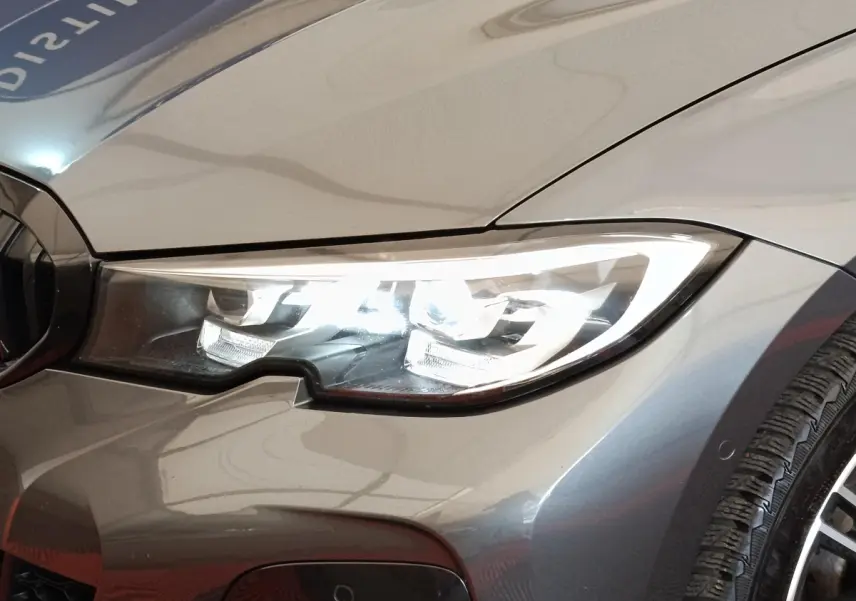 Gros plan sur le phare avant droit allumé d'une BMW Série 3 320e M Sport gris foncé, avec partie de la roue visible.