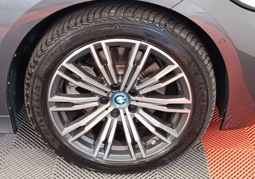 Gros plan sur la roue avant droite avec jante alliage et logo BMW, carrosserie gris foncé visible en arrière-plan.