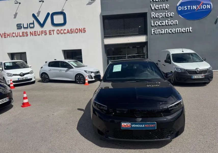 Opel Corsa 1.2 Turbo Hybrid noir vue de face, stationnée devant un garage avec panneaux de vente visibles.