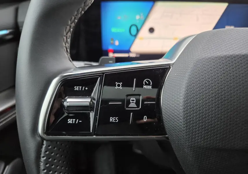 Gros plan sur les commandes gauche du volant noir du Renault Austral 2025 avec écran numérique flou en arrière-plan.