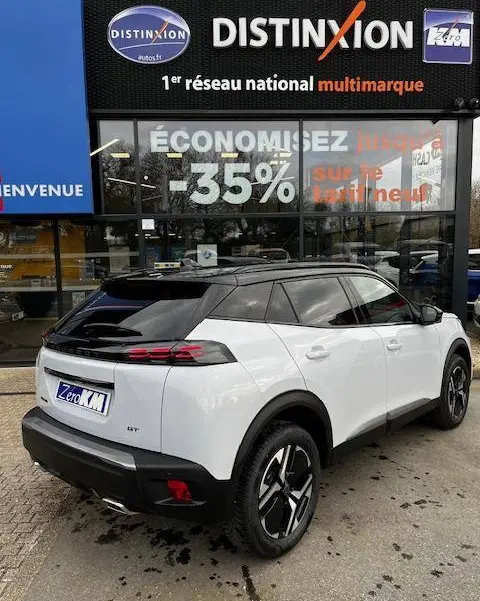 Vue 3/4 arrière droite d'un Peugeot 2008 GT blanc avec toit noir et jantes distinctives, stationné devant un concessionnaire multimarque.