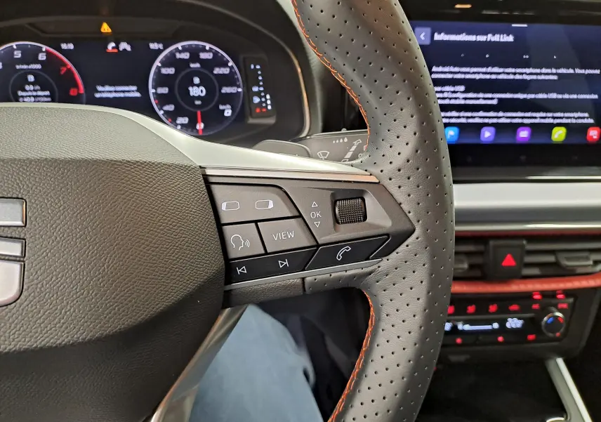 Gros plan sur le volant perforé avec surpiqûres orange du SEAT Arona 2024, tableau de bord numérique et écran tactile en arrière-plan.